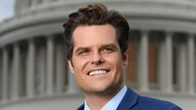 Matt Gaetz smiling in a blue suit and pink tie in front of the US Capitol dome