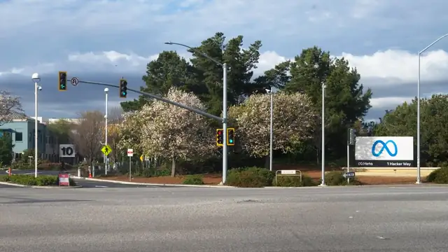 The white Meta sign at the 1 Hacker Way entrance to the company's headquarters in Menlo Park beside a busy intersection with traffic lights and blooming trees