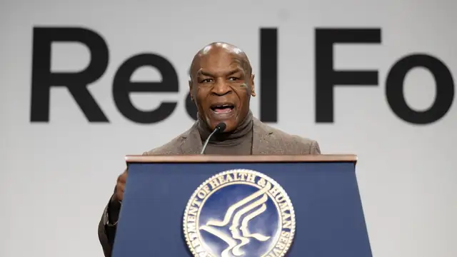 Mike Tyson in a brown blazer speaks at a blue podium in front of a large white sign with black lettering