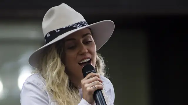 Miley Cyrus singing into a microphone under a cream wide-brimmed hat with a bandana band