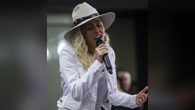 Miley Cyrus singing into a microphone in a white denim jacket and a wide-brimmed cream hat