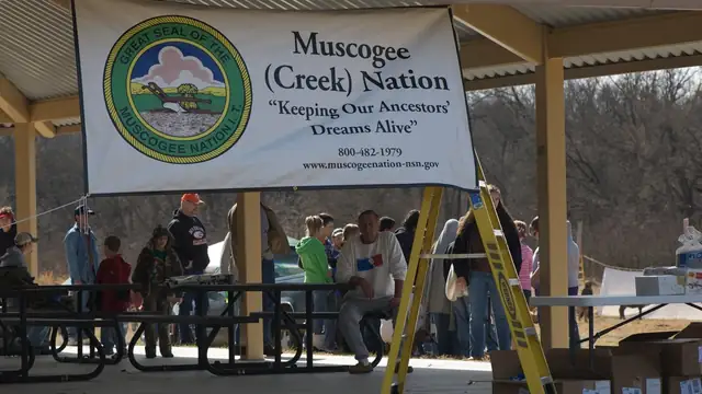 A Muscogee Creek Nation banner hangs under a covered pavilion where people gather at picnic tables