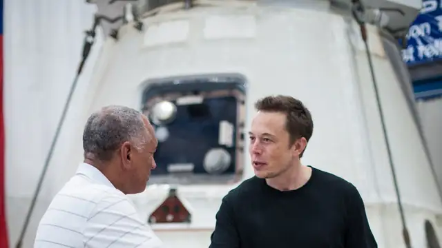 Elon Musk in a black T-shirt talks with then-NASA Administrator Charles Bolden beside a SpaceX Dragon capsule