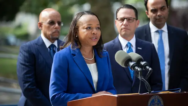 Kristen Clarke speaking at a podium with officials in dark suits standing behind her at a Justice Department announcement