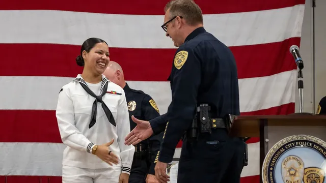 Navy Hospital Corpsman Navaeh Funderburk in white dress uniform shakes hands with a National City police officer beside an American flag