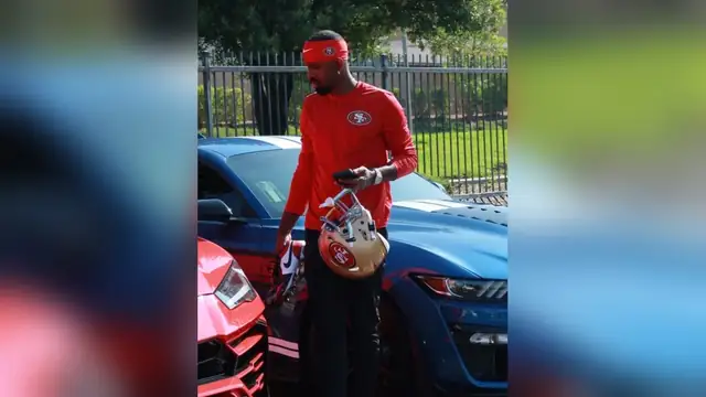 Daejon Love in a red 49ers headband and long-sleeve shirt holding a phone and a 49ers helmet beside a blue sports car