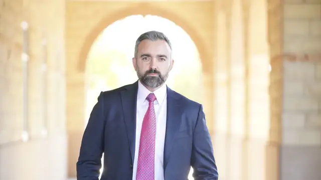 New Mexico Attorney General Raúl Torrez in a navy suit and pink tie standing in a stone archway