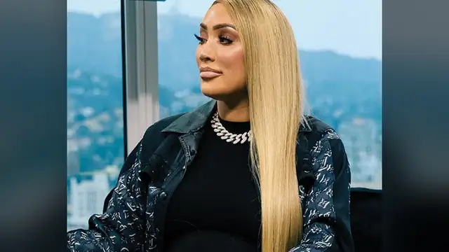 Nikki Mudarris with long straight blonde hair wearing a chunky diamond chain necklace and a patterned black jacket seated in profile in front of a hillside city view
