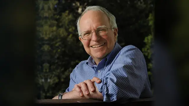 Smiling economist Robert E. Hall in glasses and a blue striped shirt leaning on a wooden rail outdoors
