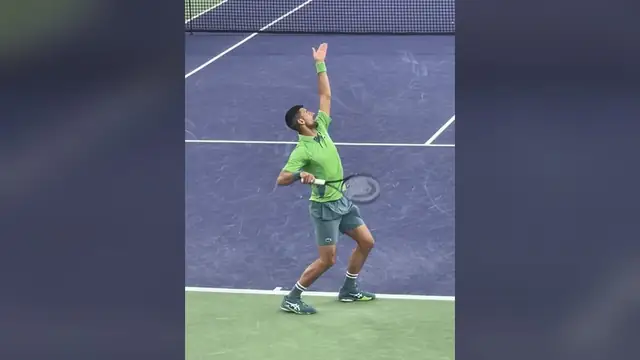 Novak Djokovic in a green shirt reaches up to serve on a blue hard court at Indian Wells
