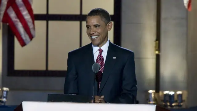 Barack Obama in a dark suit and red striped tie smiling broadly at a podium on a stage with American flags behind him