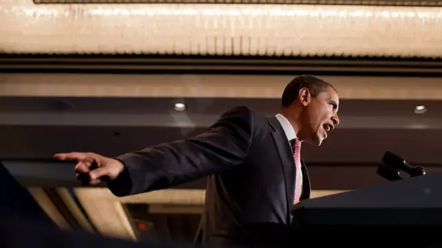 Barack Obama in a dark suit and patterned tie stretches out his arm and points as he speaks at a campaign rally lectern under chandelier lights