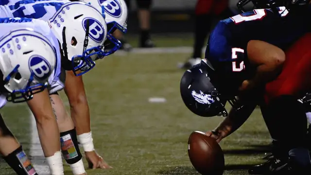 Linemen in white helmets with a blue B logo crouch across from an opposing center in a dark helmet who grips the football before the snap at a night game