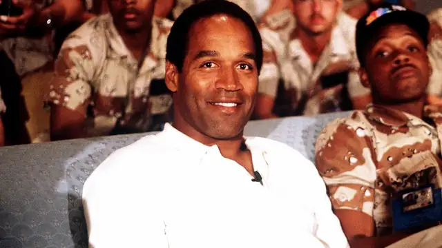 O.J. Simpson smiling in a white polo shirt while seated on a couch with US service members in 1990