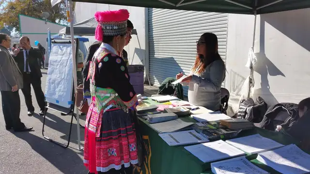 A woman in a traditional Hmong costume with a red hat stands at an information table during a Hmong New Year event