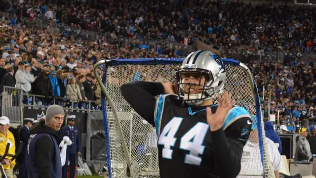 Carolina Panthers long snapper JJ Jansen in a black No. 44 jersey and silver helmet raises his hand on the sideline in front of a practice net as fans fill the stands