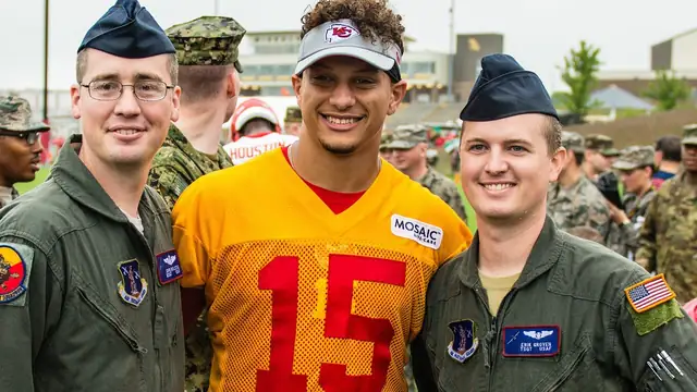 Patrick Mahomes in a gold No. 15 Chiefs practice jersey and visor smiles while posing with Air National Guard airmen at Chiefs training camp