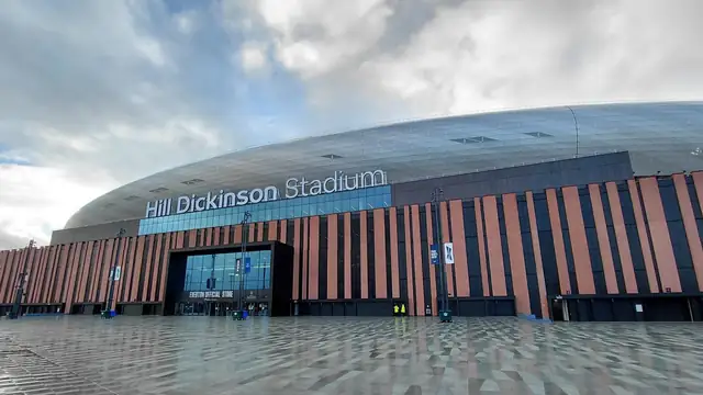 The curved silver roof and lettered sign of Hill Dickinson Stadium in Liverpool rise above a wide wet plaza under a cloudy sky