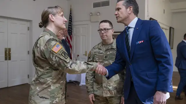 Pete Hegseth in a blue suit shaking hands with a uniformed soldier in an indoor hall with flags