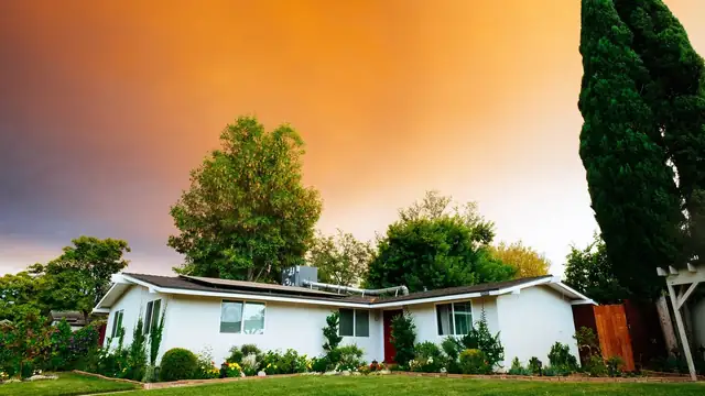 A white single-story house with a green lawn and tall trees under an orange evening sky