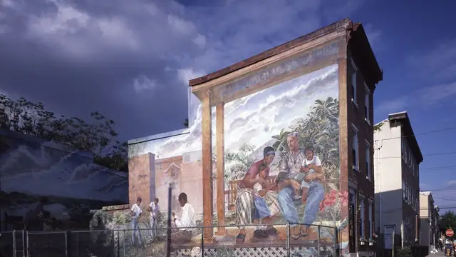 A large painted mural of a family reading together on a porch covers the side of a brick row house in a Philadelphia neighborhood