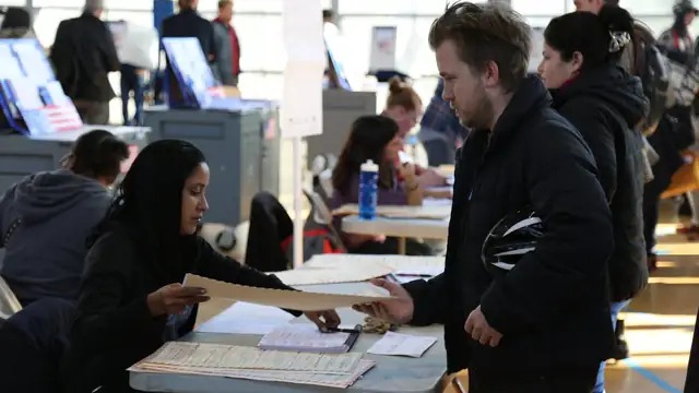 A poll worker hands a paper ballot across a folding table to a young man holding a bike helmet inside a busy gym used as a polling place