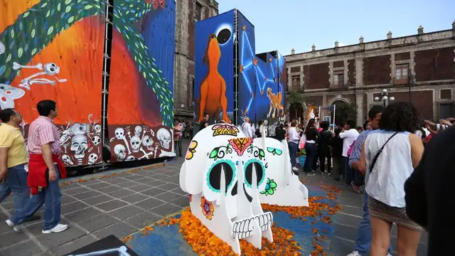 A giant painted sugar skull sits on a bed of orange marigolds as families stroll past at a Day of the Dead celebration