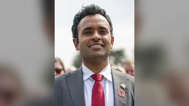 Vivek Ramaswamy smiles in a gray suit and red tie with a small red pin on his lapel and a crowd blurred behind him