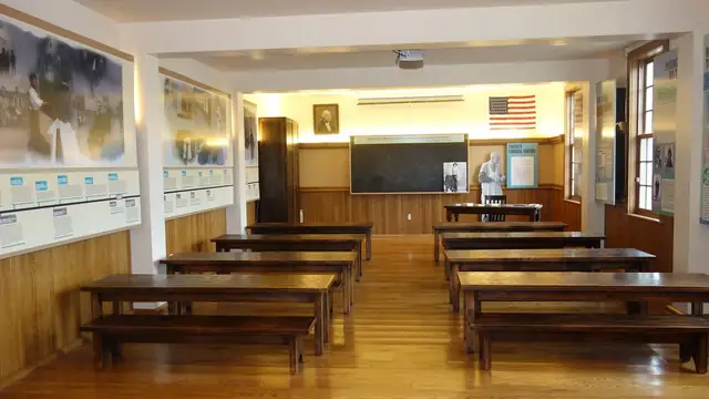 A tidy schoolroom with rows of wooden desks and benches facing a chalkboard and an American flag