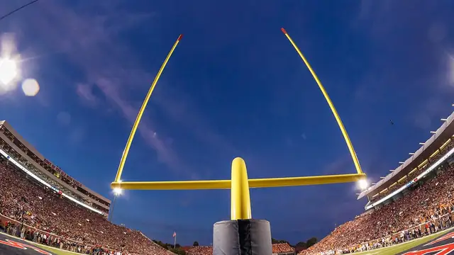 Yellow football goal posts rise into an evening sky above a packed college stadium