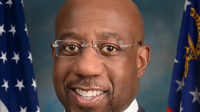 Official Senate portrait of Sen. Raphael Warnock smiling in a dark suit and striped tie in front of flags