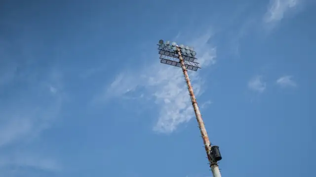A tall stadium light tower rises into a blue sky with thin white clouds