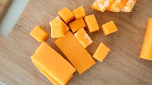 Yellow cubes and blocks of cheddar cheese sit on a wooden cutting board
