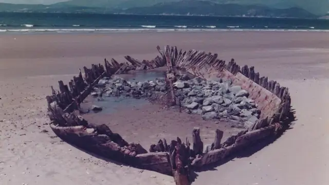 The weathered wooden ribs of an old shipwreck sit half buried in the sand on a wide beach with the ocean and hills behind it
