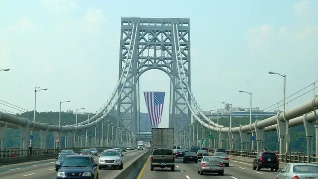 Cars and a box truck head west on the upper deck of the George Washington Bridge toward a tower hung with a giant American flag