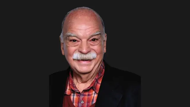 Richard Riehle smiling with a white mustache in a red plaid shirt and dark blazer