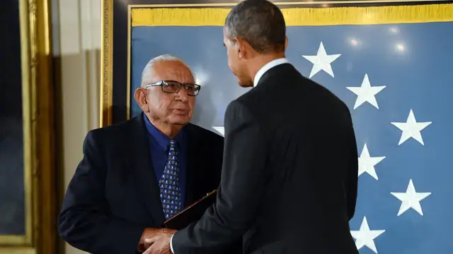 Alfonzo Lara shaking hands with President Barack Obama at the White House Medal of Honor ceremony in front of a blue flag with white stars