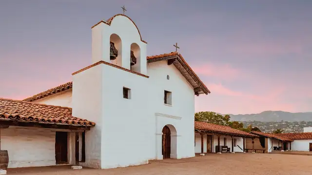 The white adobe chapel of El Presidio de Santa Barbara with its bell tower and red tile roofs under a pink evening sky