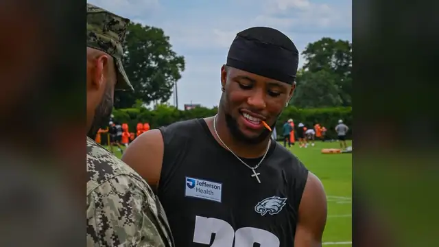 Saquon Barkley smiling in a black Eagles No. 26 practice jersey as he signs a football for a service member at training camp in 2025