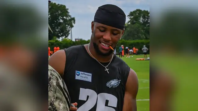 Saquon Barkley smiling in a black Eagles No. 26 practice jersey while signing a football for a service member at training camp