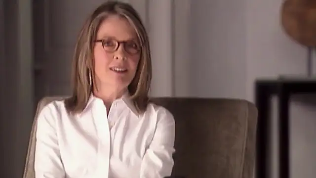 Diane Keaton in glasses and a white shirt smiling in an armchair
