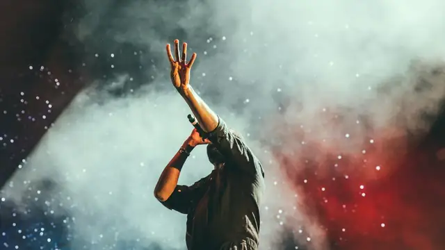 A Black performer on stage holding a microphone with one arm raised under red and blue concert lights