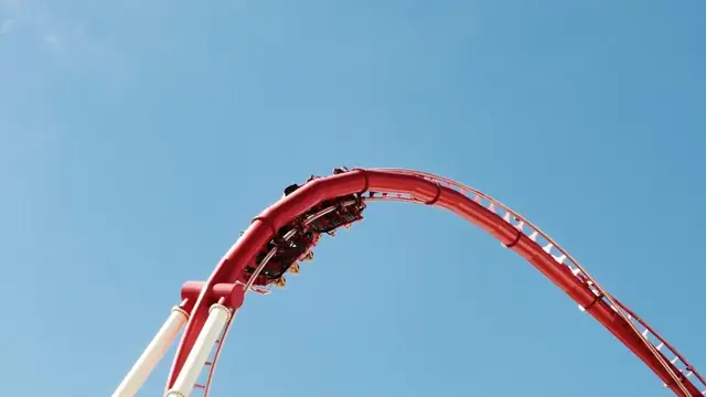 Riders in a red coaster car at the top of a loop against a bright blue sky