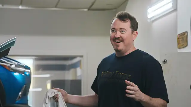 Shane Gillis smiling in a navy Valley Forge T-shirt in the auto shop in a still from the Netflix series Tires