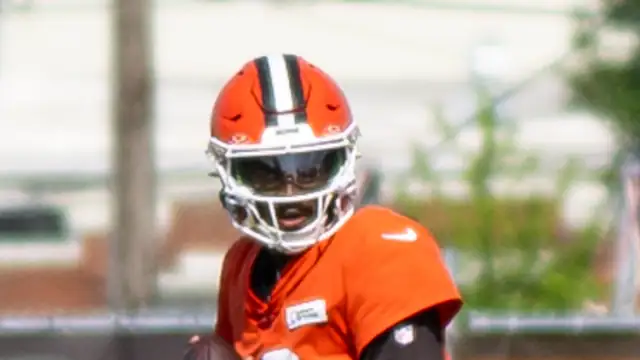 Shedeur Sanders in an orange Browns helmet and practice jersey holding a football at training camp