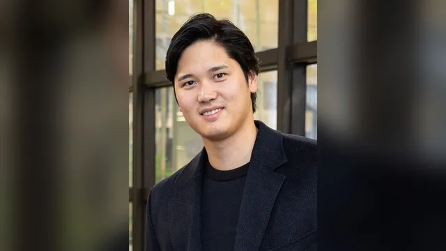 Shohei Ohtani in a dark suit and striped tie smiles while standing outdoors against a wooden wall