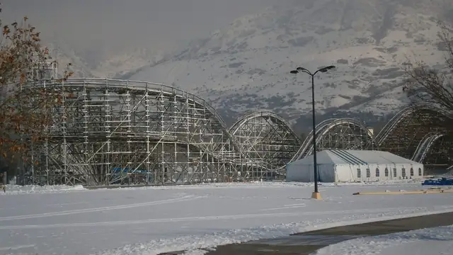 A wooden roller coaster standing idle in a snowy amusement park below a mountain