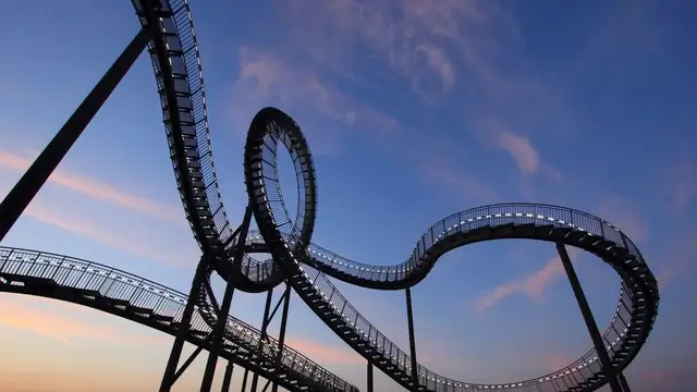 A twisting steel track sculpture shaped like a roller coaster against a pink and blue dusk sky