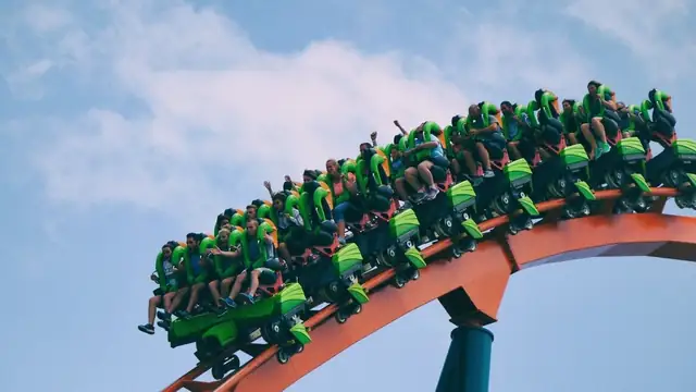 A roller coaster train climbing an orange steel track against a pale blue sky