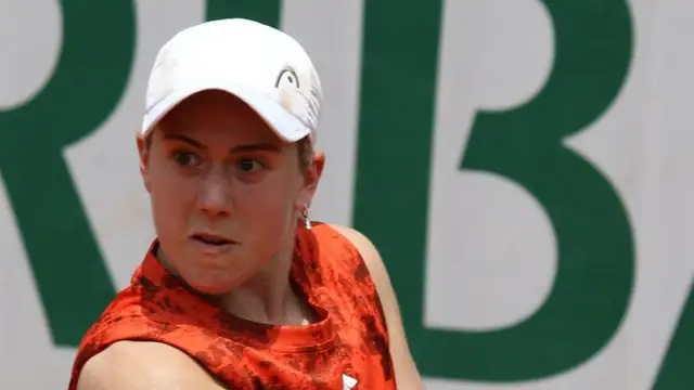 Sonay Kartal in a white cap and orange sleeveless top turns to hit a backhand in front of a green and white wall at the 2023 French Open